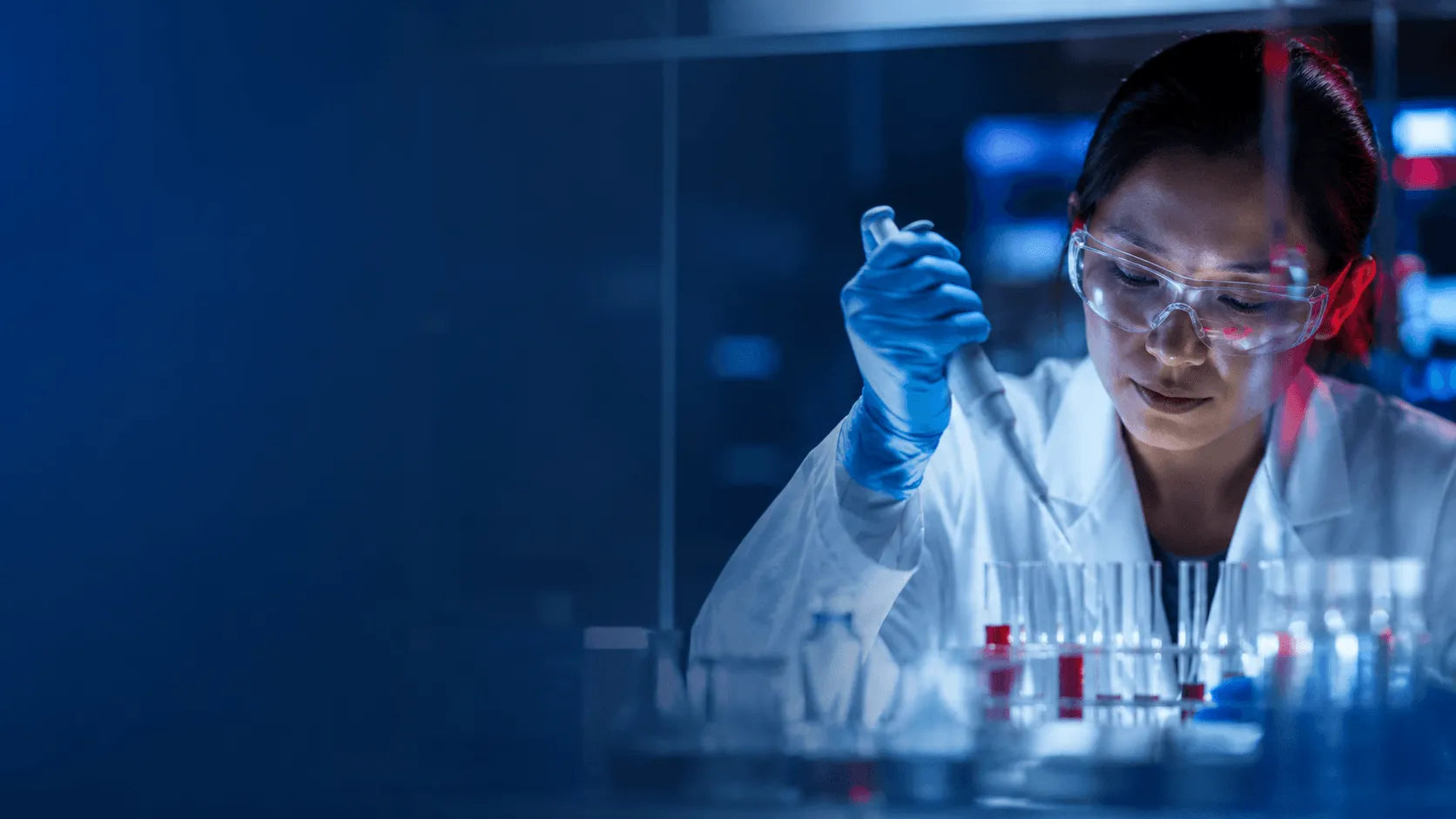 A female scientist in a white lab coat, clear safety goggles, and blue gloves holds a test tube under a glass incubator in a dark laboratory setting.